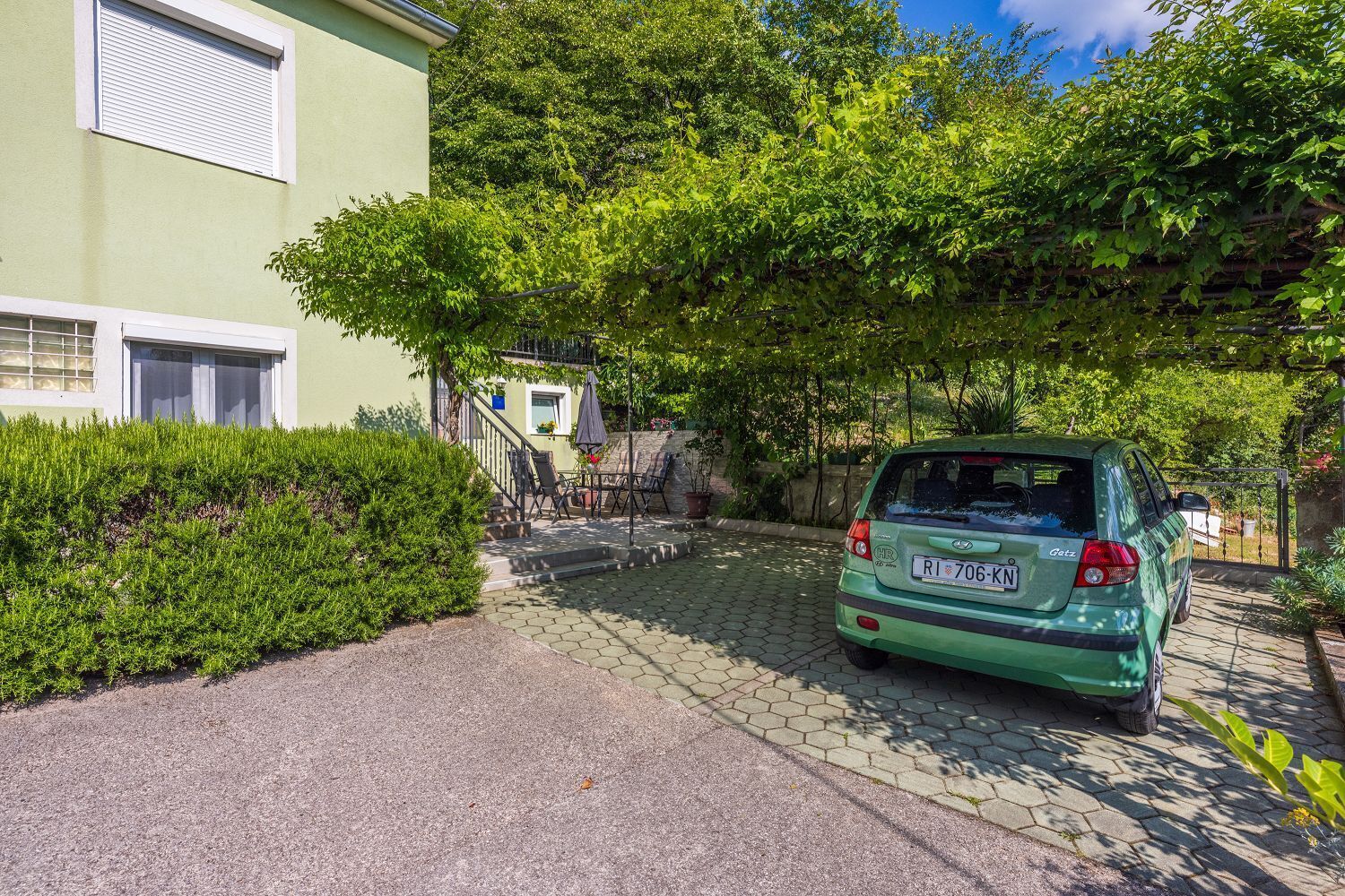 A green car parked in front of a house with a paved driveway and a covered outdoor seating area.