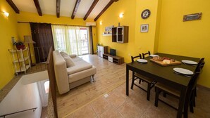Una habitación luminosa de color amarillo con techo de madera, un sofá, una mesa de comedor para dos personas y una pequeña zona de cocina.