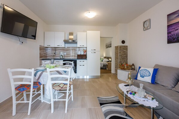 A modern living room with a sofa, a dining area with chairs and a table, a kitchen with cabinets and a stove, and a TV mounted on the wall.