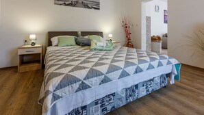 A neatly made bed with a geometric patterned bedspread, a nightstand with a lamp, and a wall-mounted picture of a pier. - Renovated flat with sea view, fitness room, baby cot, barbecue area (Crikvenica)