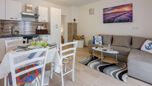 A modern living room with a grey sofa, a dining table set for four, and a kitchen area with white cabinets and a stainless steel range hood. - Renovated flat with sea view, fitness room, baby cot, barbecue area (Crikvenica)