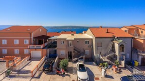 Un quartier résidentiel situé sur la côte, avec des bâtiments aux toits de tuiles rouges, une allée pavée, des véhicules garés et une vue dégagée sur la mer.
