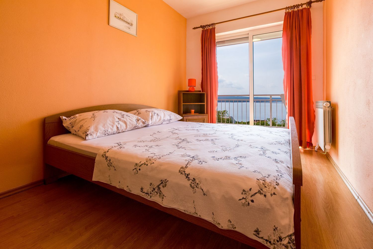 Ein Schlafzimmer mit Bett, Nachttisch, einem Fenster mit Vorhängen und Blick ins Freie.