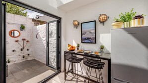 Una cocina moderna con puerta de cristal que da a un patio, un refrigerador, dos taburetes de bar, una mesa de comedor con un frutero y un cuadro en la pared.