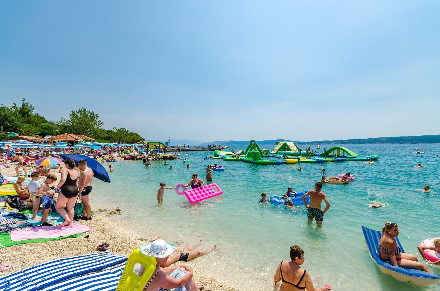 Una playa concurrida donde gente nada y juega en el agua, con diversas actividades como estructuras inflables y tumbonas.