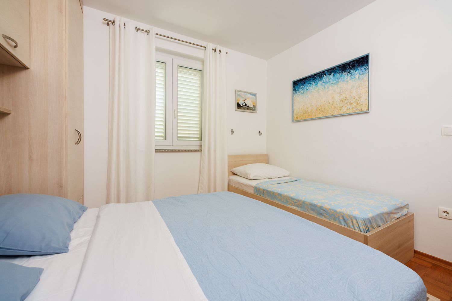 Une chambre à coucher avec un lit, une fenêtre avec des rideaux, une table de chevet, un tableau accroché au mur et une armoire en bois.