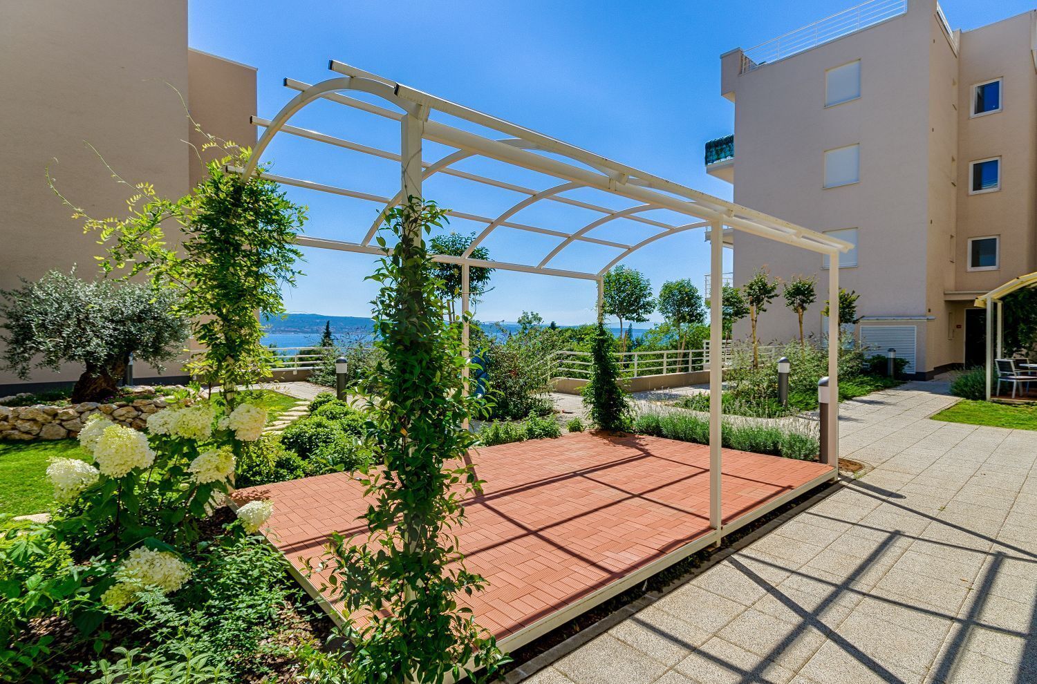 A covered outdoor area with a pergola, surrounded by greenery and a view of the sea.