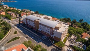 An aerial view of a coastal hotel with a large building, a road with a pedestrian crossing, and a view of the sea and distant mountains. - Modern flat only 90 metres to the beach with sea view, underground car park, air conditioning (Crikvenica)