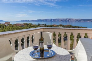 Eine Terrasse mit Tisch für zwei Personen, einer Weinflasche und zwei Gläsern auf einem Tablett. Die Terrasse bietet Blick auf ein Gewässer und entfernte Berge.