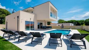 A modern two-story house with a swimming pool, outdoor seating, and a stone wall.