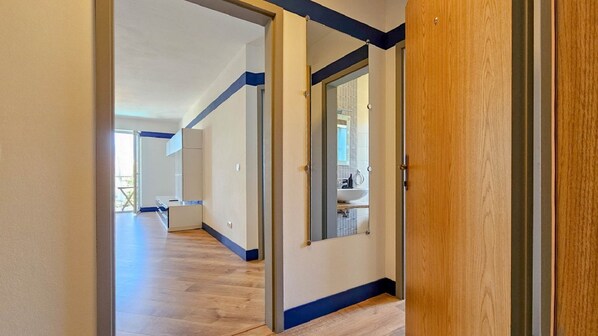 Un pasillo con suelo de madera, pared blanca y puerta que da a un baño con espejo y lavamanos.