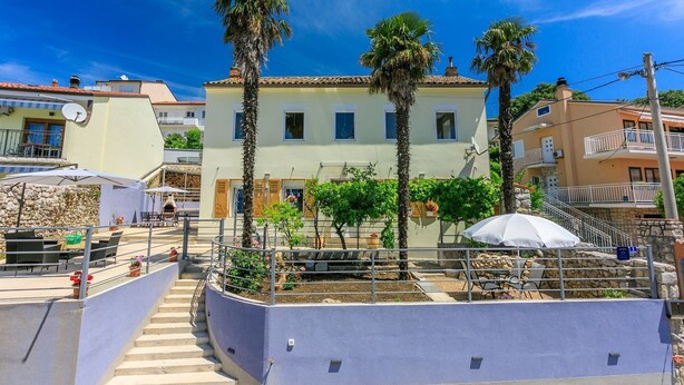 A coastal building with a terrace, palm trees, and outdoor seating.