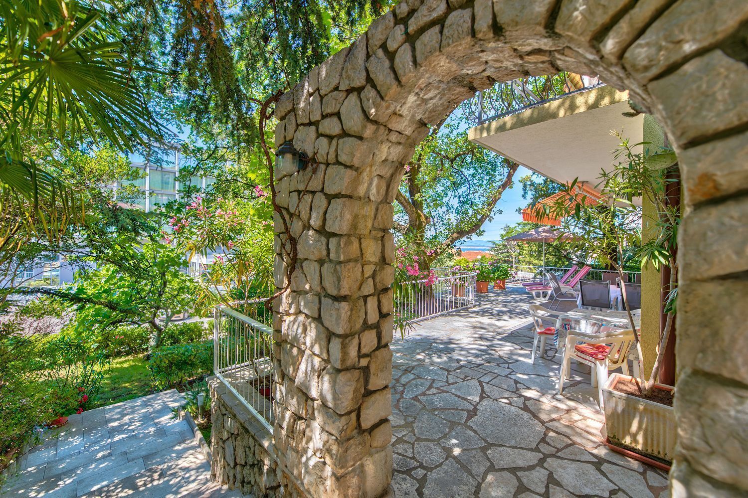 A stone pathway leading to a covered outdoor seating area with tables and chairs, surrounded by greenery and a stone wall.
