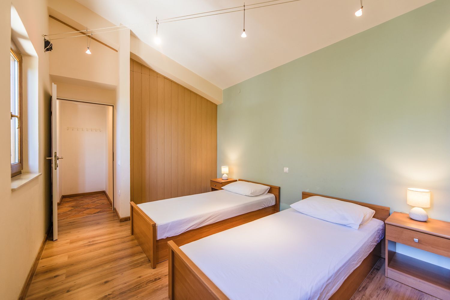 A room with two wooden beds, a wooden wardrobe, a small bedside table with a lamp, and a door leading to another room.
