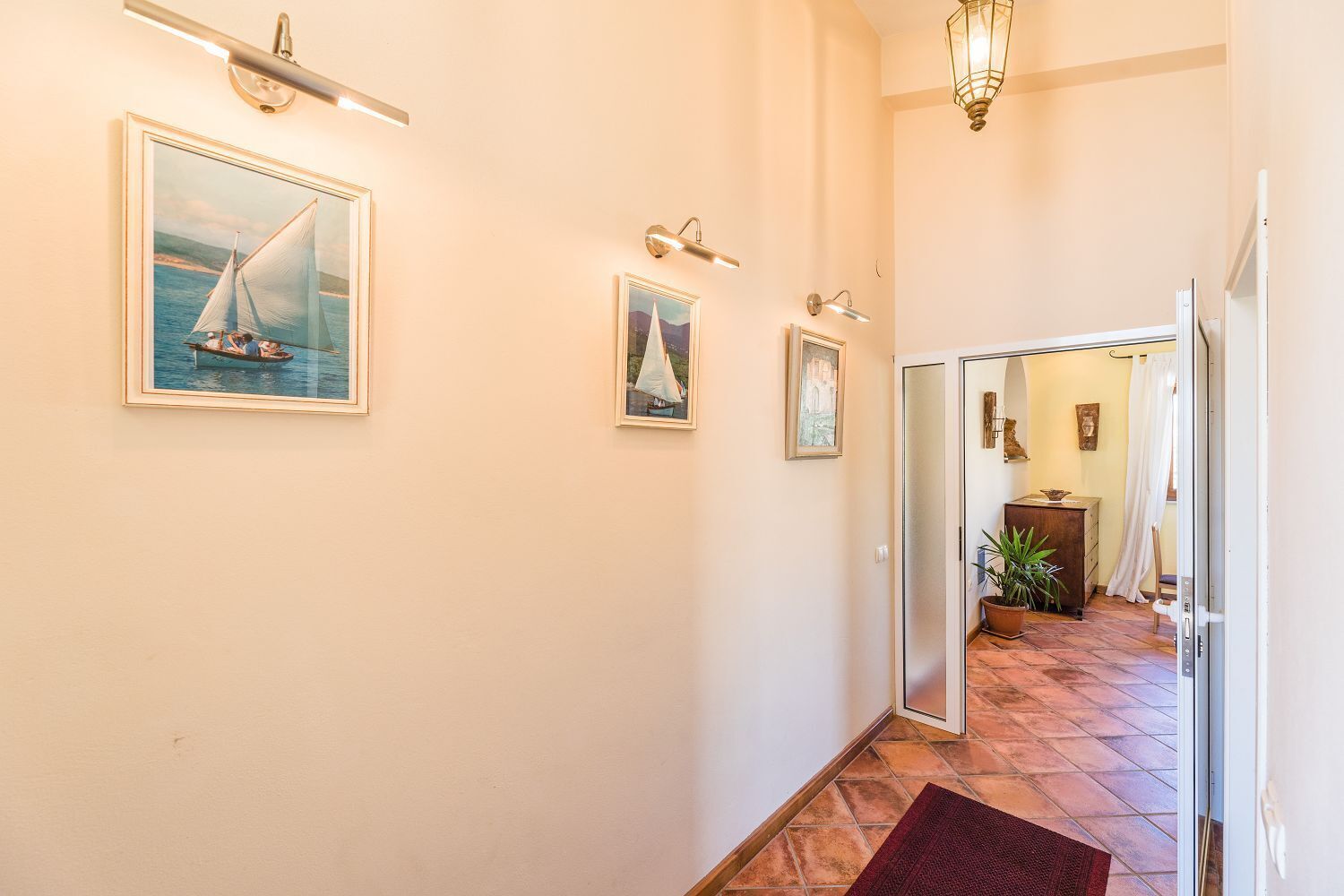 Een gang met aan de wanden gemaritimeerde schilderijen in lijsten, een betegelde vloer en een deur naar een andere kamer.