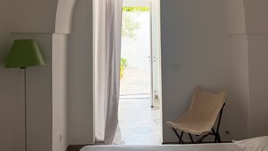 A bedroom with a bed, a chair, a lamp, and an arched doorway leading to an outdoor area.