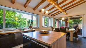 Een moderne keuken met een groot eiland, houten balken en grote ramen met uitzicht op groen.