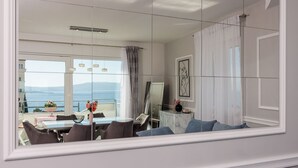 Una sala moderna con un ventanal grande con vista al mar, una zona de comedor con mesa y sillas, y una consola de mármol con objetos decorativos.