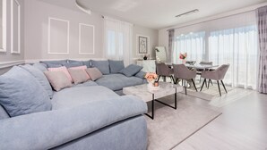 Una sala moderna con un sofá seccional, un comedor con mesa de vidrio y ventanas grandes con cortinas de gasa.