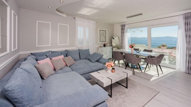Una sala de estar moderna con un sofá seccional gris, un comedor con mesa y sillas, y un ventanal con vista al mar.