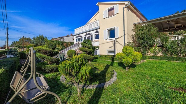 Une maison à deux étages avec un jardin bien entretenu, un escalier visible et une chaise en métal au premier plan.