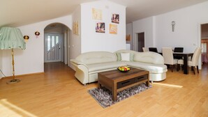 Un salon avec un canapé d’angle beige, une table basse en bois sur laquelle sont disposés des fruits, une lampe, et un coin repas avec une table et des chaises.