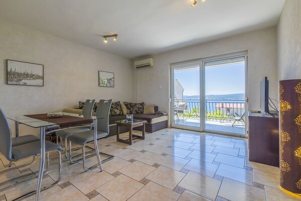 A modern living room with a dining area, large sliding glass doors to a balcony, and a flat-screen TV on a wooden cabinet.