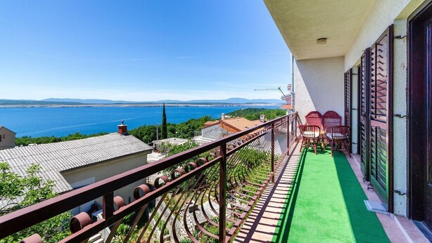 Un balcone con vista sul mare, un tappetino verde e sedie rosse.