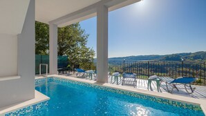 A pool area with clear blue water, surrounded by lounge chairs and a view of a hilly landscape.