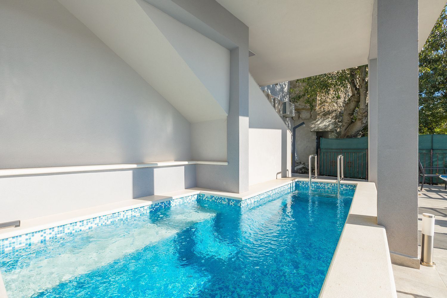 Une piscine extérieure moderne, avec une eau d’un bleu limpide, un mur en pierre et un escalier en métal.
