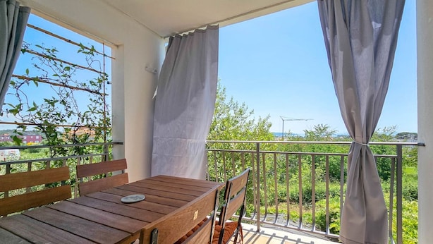 Ein Balkon mit Holztisch und -stühlen, Vorhängen und Blick auf Grünflächen und Gebäude.