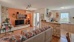 Ein modernes Wohnzimmer mit Sofa, Fernseher und Unterhaltungseinheit. Ein Küchenbereich mit Steininsel und Schränken ist zu sehen.