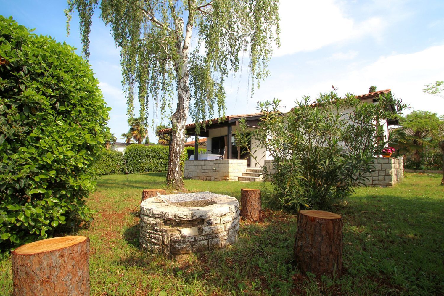 A stone well, a wooden bench, and a stone wall in a garden with a house in the background.