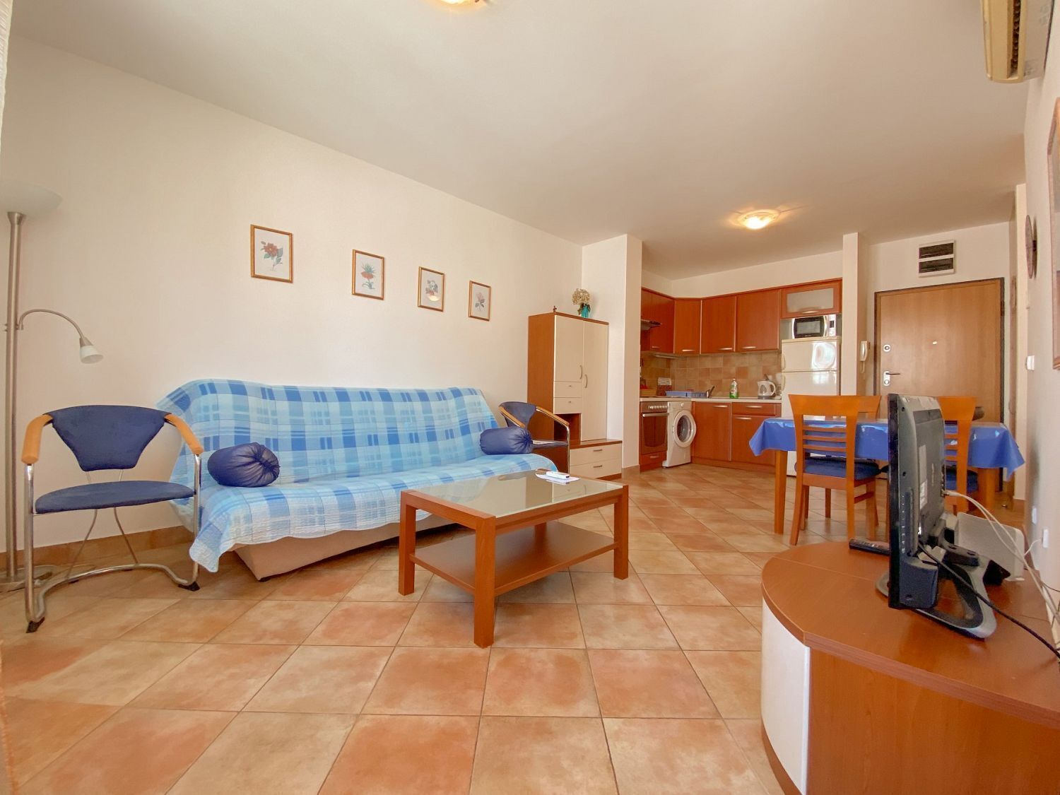 A living room with a sofa, a coffee table, and a TV. There is a kitchen in the background with a microwave and a washing machine.