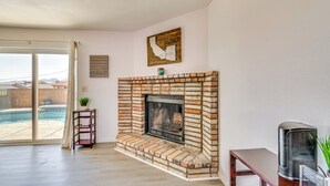 Ein Wohnzimmer mit Kamin aus Ziegelsteinen, einem Fernseher auf einem Lowboard und Blick auf einen Swimmingpool durch eine Glastür.
