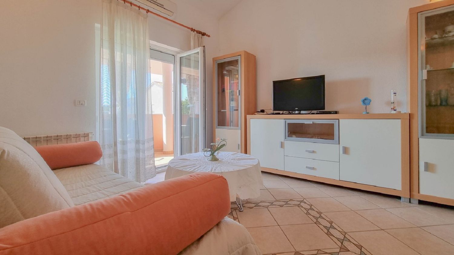 A living room with a sofa, a television on a cabinet, and a glass door leading outside.