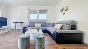 Ein modernes Wohnzimmer mit einer Eckcouch, zwei Hocker, einem Couchtisch und einem Fernseher auf einem weißen Schrank.