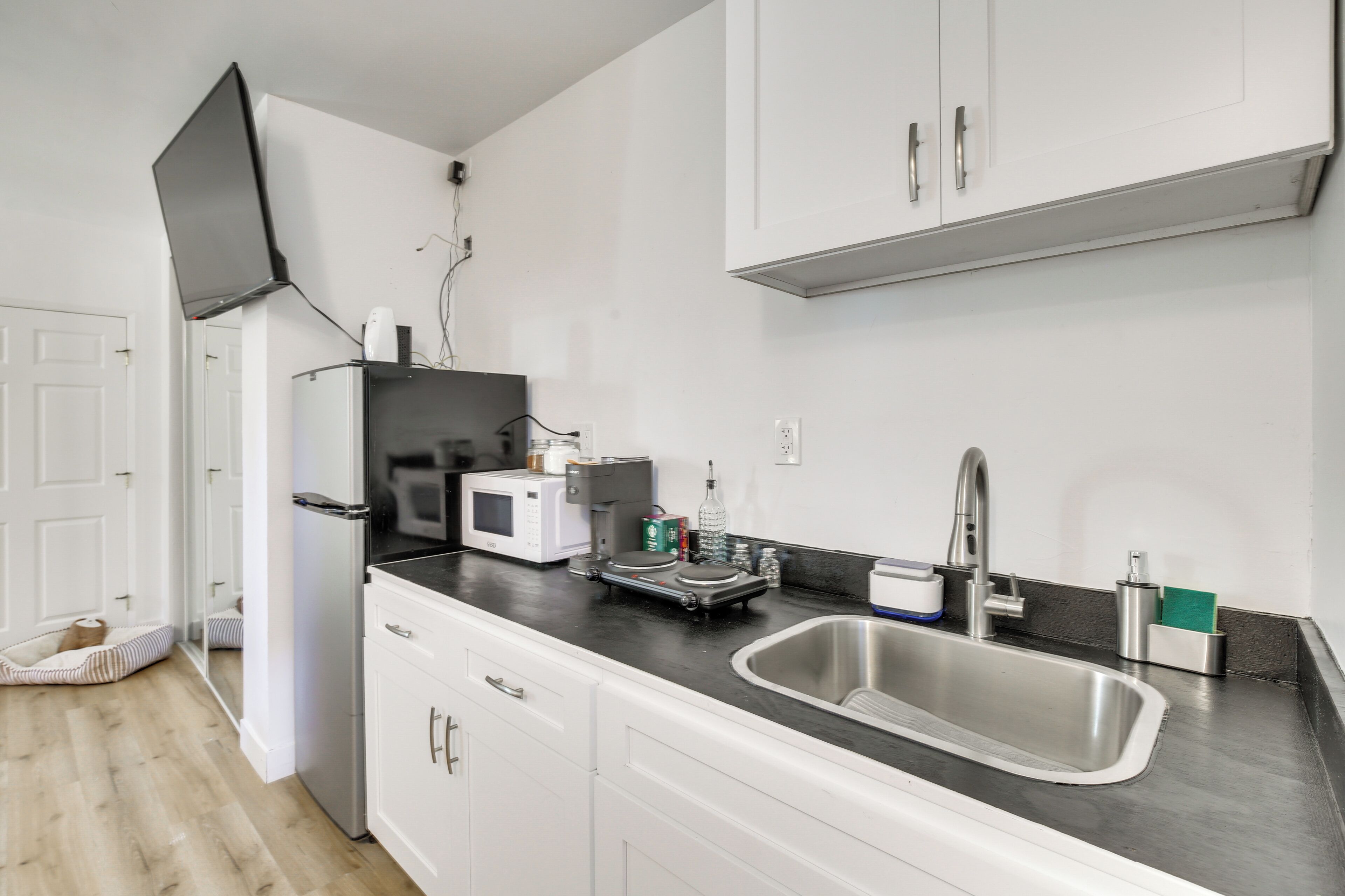 A modern kitchen with white cabinets, a stainless steel sink, a microwave, and a flat-screen TV mounted on the wall.