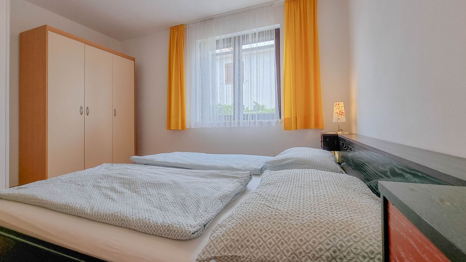 A bedroom with a bed, a wardrobe, a window with yellow curtains, and a bedside lamp.
