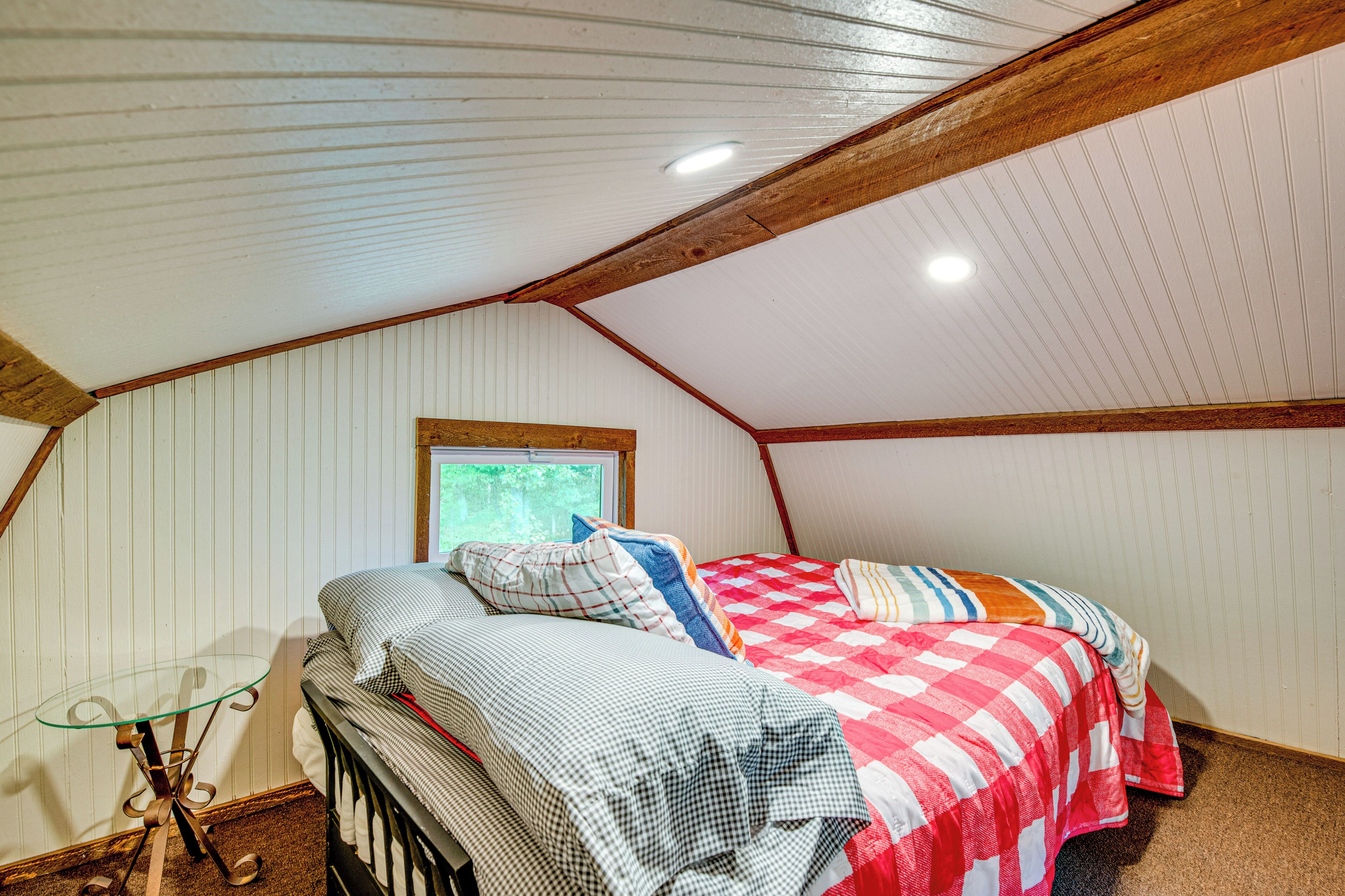 Un dormitorio con techo inclinado, una ventana, una cama con colcha de cuadros y una pequeña mesa con lámpara.