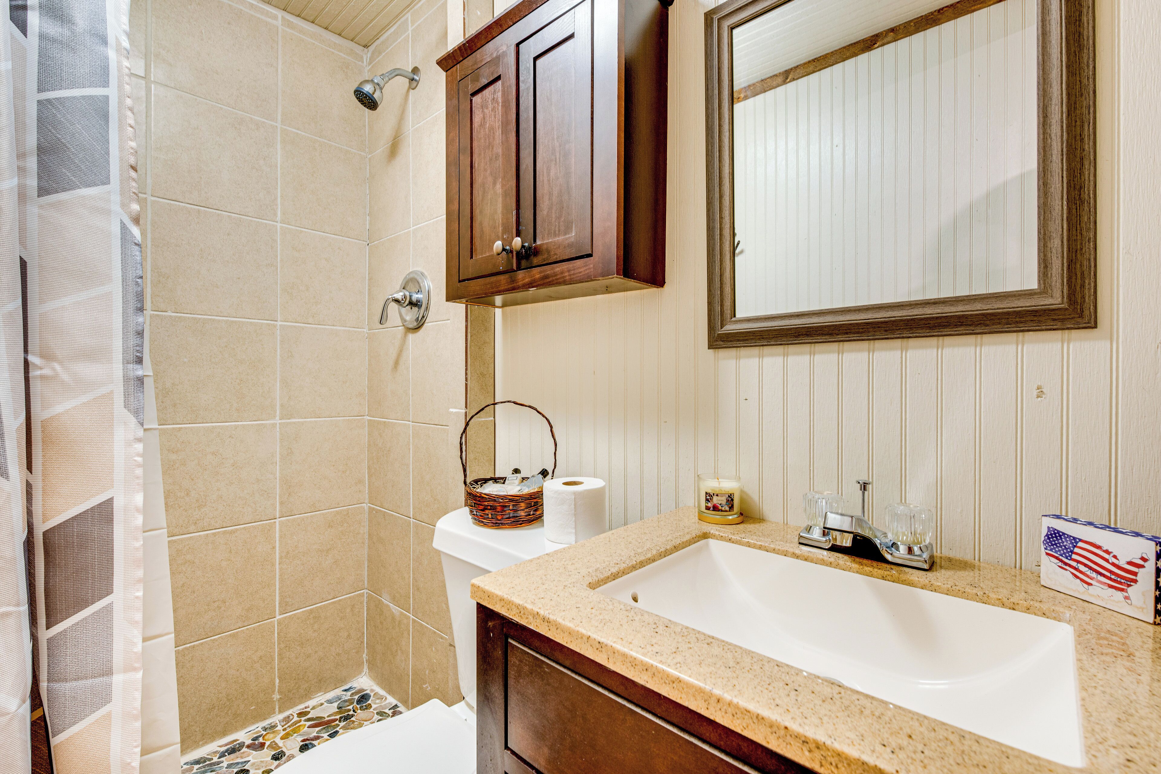 Un baño con ducha de azulejos color beige, un armario de madera, un lavabo con espejo y una pequeña decoración con la bandera estadounidense.