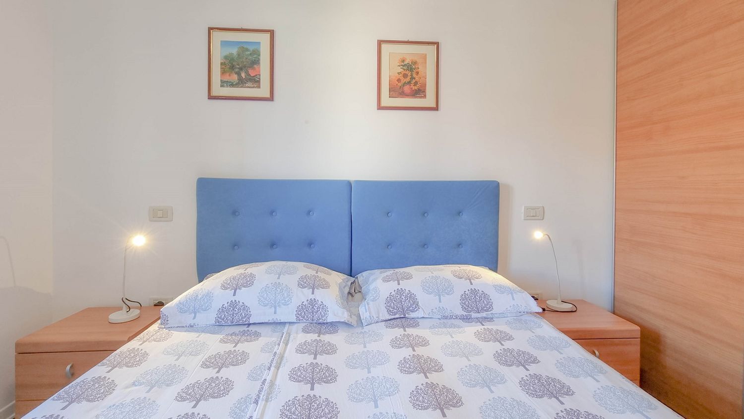 A bedroom with a blue headboard, two bedside tables with lamps, a bed with a tree-patterned duvet, and two framed pictures on the wall.