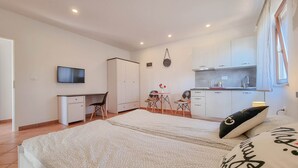 A bedroom with a bed, a desk, a chair, a small table with flowers, and a kitchenette.