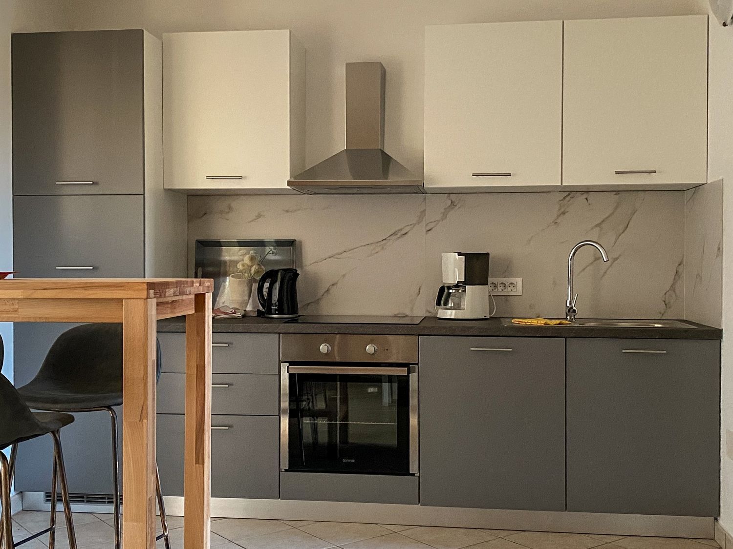 A modern kitchen with grey cabinets, a stainless steel range hood, a built-in oven, a coffee maker, and a sink with a faucet.