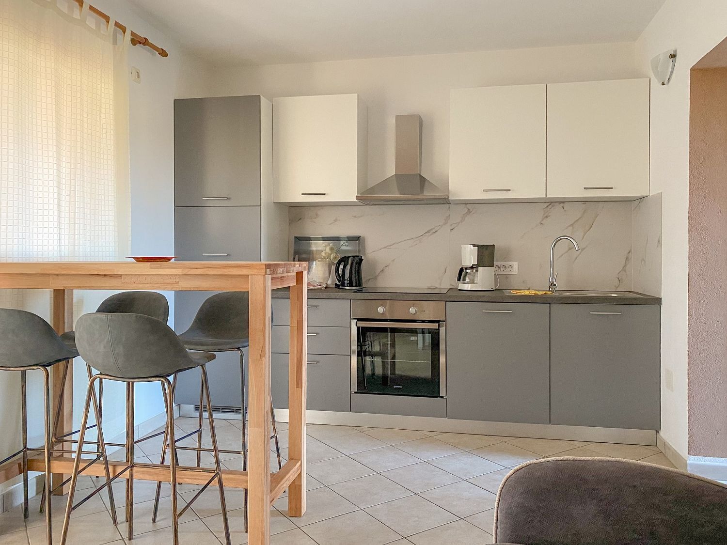 Eine moderne Küche mit grauen Schränken, einer Edelstahl-Dunstabzugshaube, einer Kaffeemaschine und einem eingebauten Backofen. Ein Holztisch wird von vier Stühlen begleitet.