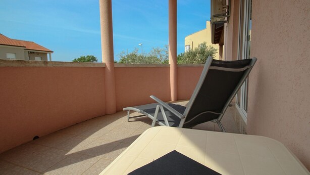 Ein Balkon mit einem Stuhl und einem kleinen Tisch, eine beige Wand und ein klarer, blauer Himmel im Hintergrund.