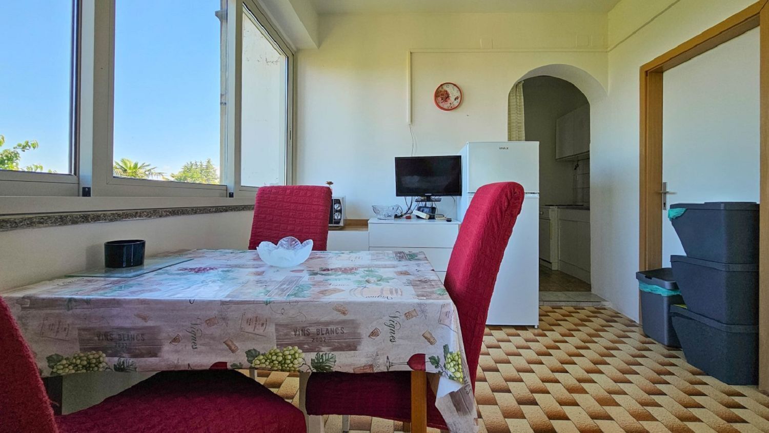 Un coin repas avec une table dressée pour deux personnes, un téléviseur et une cuisine visible à travers une arche.