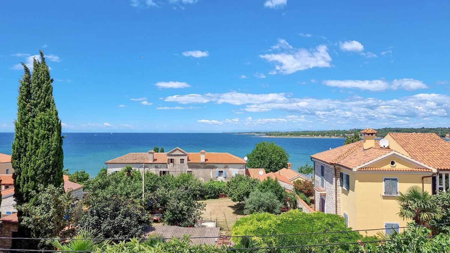 A coastal village with traditional architecture, including terracotta-roofed houses and a prominent cypress tree.