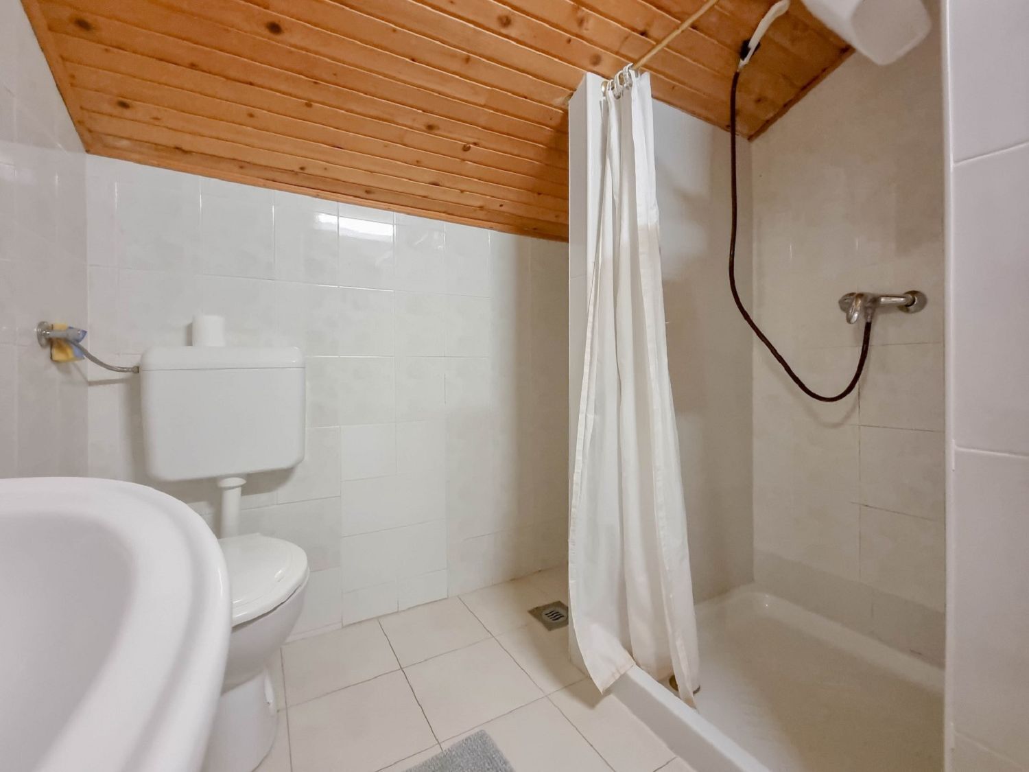 Un baño con techo de madera, azulejos blancos, un inodoro, una bañera y una cortina de ducha.