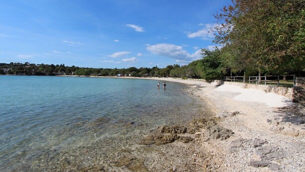 Una playa con agua cristalina, costa rocosa y un camino bordeado de árboles.